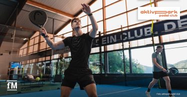 Zwei Männer spielen Padel auf einem Indoor-Court. Im Vordergrund bereitet sich ein Spieler mit Schläger auf den Ball vor.