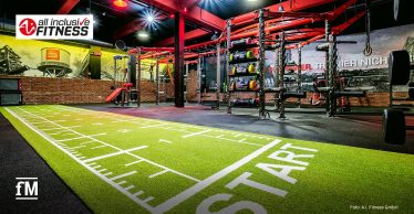 Ein weitläufiger Fitnessbereich mit leuchtend grüner Kunstrasenbahn, auf der das Wort 'Start' steht. Dahinter befinden sich strukturierte Trainingsgeräte, Hanteln und eine Wand mit Sportmotiven. Der Raum ist rot und schwarz gestaltet.