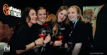Im Vordergrund stehen vier Frauen mit Gläsern vor einem dunklen Messestand. Links im Hintergrund leuchtet der Schriftzug „all inclusive FITNESS“