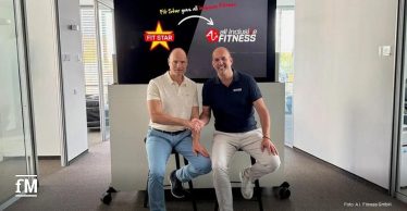 Zwei Männer sitzen nebeneinander und schütteln sich die Hand. Hinter ihnen zeigt ein Bildschirm die Logos von 'Fit Star' und 'all inclusive FITNESS' mit dem Text 'Fit Star goes all inclusive Fitness'.