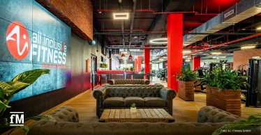 Moderner Fitnessstudio-Eingangsbereich mit mehreren grauen Chesterfield-Sofas, Holztischen, großen Pflanzenkübeln und roten Säulen; links ein großer Bildschirm mit dem Logo 'all inclusive Fitness'.