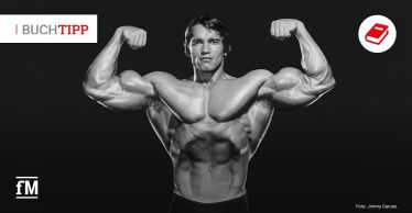 Schwarz-weißes Foto eines Bodybuilders mit erhobenen Armen vor schwarzem Hintergrund, links oben steht „BUCHTIPP“.