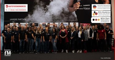 Eine große Gruppe von Menschen steht lächelnd vor einer Sponsorenwand mit Logos verschiedener Partner. Alle tragen dunkle Kleidung und Namensschilder, im Hintergrund ist ein Sportmotiv mit Rauchwolke zu sehen