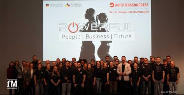 Eine große Gruppe von Menschen steht lächelnd vor dem Logo des Aufstiegskongress 2025. Es zeigt zwei Menschen, die Rücken an Rücken stehen, mit dem Motto 'Powerful. People | Business | Future'.