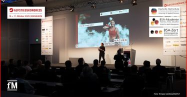 Auf der Bühne des Aufstiegskongresses 2025 steht eine Rednerin vor einer großen Leinwand und spricht zu einem Publikum. Auf der Leinwand ist die Präsentation von Prof. Dr. Sarah Kobel zum Thema 'Fitnesstypen next level' zu sehen.