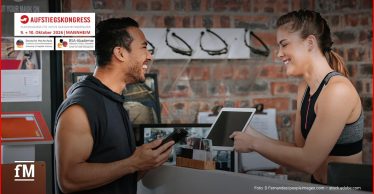 Frau mit Tablet und Mann mit Smartphone im Gespräch am Empfang eines Fitnessstudios.