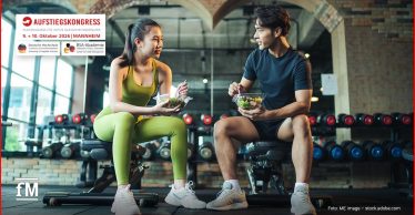 Zwei Personen sitzen mit Salatschalen auf Trainingsbänken in einem Fitnessstudio
