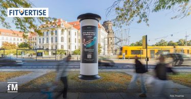 Litfaßsäule mit Werbung für ein Fitnessstudio, an einer Straße mit vorbeifahrender Straßenbahn und Passanten, im Stadtzentrum.