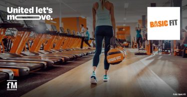 Frau mit Basic-Fit-Sporttasche geht zwischen Laufbändern durch ein Fitnessstudio