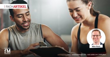 Ein Trainer mit zeigt einer jungen Frau ein Klemmbrett. Beide lächeln und sitzen in einem Fitnessstudio.