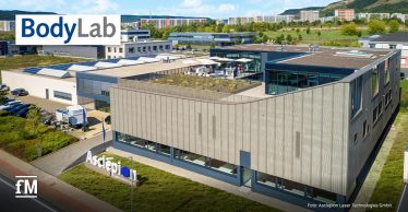 Luftaufnahme des modernen Asclepion-Firmensitzes mit großer Fensterfront und begrüntem Dach. Menschen versammeln sich auf der Dachterrasse. Im Bild sind die Logos „BodyLab“ und „fM“ eingeblendet