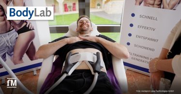 Ein Mann liegt auf einer Liege und lächelt. Zwei Bodyforming-Applikatoren sind an seinem Bauch befestigt. Im Hintergrund sind Poster mit Fitnessmotiven und Informationen zur Behandlung zu sehen.