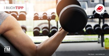 Eine Person in einem Fitnessstudio führt auf einer Trainingsbank eine Bizepsübung mit einer Kurzhantel aus. Im Hintergrund sind viele weitere Hanteln zu sehen.