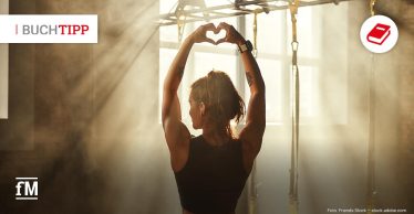 Sportlerin im Fitnessstudio formt mit den Händen ein Herz über dem Kopf. Sonnenstrahlen fallen durch die Fenster und erzeugen eine motivierende, positive Stimmung.