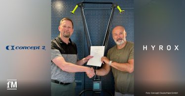 Zwei Männer (links Glenn Dwyer und Christian Toetzke) stehen in der Bildmitte vor einem Fitnessgerät (SkiErg), reichen sich die Hand und halten gemeinsam ein unterzeichnetes Dokument. Beide lächeln in die Kamera. Links ist das Logo von Concept2 und rechts das von HYROX vor unscharfem Hintergrund.