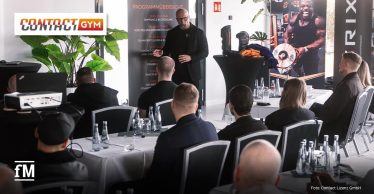 Im Vordergrund sitzen Teilnehmende an gedeckten Konferenztischen. In der Bildmitte spricht eine Person vor einem Aufsteller mit der Überschrift „PROGRAMMÜBERSICHT“. Rechts stehen ein Stehtisch mit Kampfsportausrüstung, ein Banner mit Athletenmotiv und ein vertikales Logo von Matrix Fitness. Im Hintergrund sind Pflanzen, Fensterflächen und weitere Sitzplätze sichtbar.
