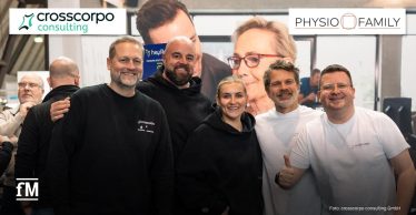 Im Vordergrund stehen fünf Personen nebeneinander auf einer Messefläche, frontal zur Kamera ausgerichtet, teilweise mit verschränkten Armen oder Daumen hoch; im Hintergrund ein großer Messestand mit der Aufschrift „crosscorpo“ und der Zeile „Partner für Therapie und Training“, weitere Personen und Standstrukturen sind seitlich und im hinteren Bereich sichtbar.