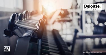 Im Vordergrund ist eine Hantelreihe auf schwarzem Rack abgebildet, Metallgriffe sind horizontal ausgerichtet; im Hintergrund ein unscharfer Fitnessraum mit Geräten und Fenstern.