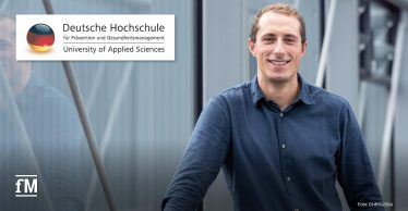 Ein Mann (Andreas Barz) im blauen Hemd steht lächelnd vor einem modernen Gebäude, links oben das Logo der Deutschen Hochschule für Prävention und Gesundheitsmanagement (DHfPG).
