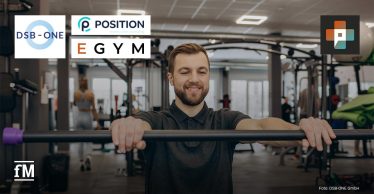 Ein Mann trainiert an einem Gerät in einem modernen Fitnessstudio und lächelt konzentriert. Im Hintergrund sind weitere Personen beim Training zu sehen. Links oben links die Logos von DSB-ONE, Positin Consulting und EGYM, rechts oben das Logo von Physio+.
