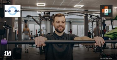 Ein Mann trainiert an einem Gerät in einem modernen Fitnessstudio und lächelt konzentriert. Im Hintergrund sind weitere Personen beim Training zu sehen. Links oben das Logo von DSB-ONE, rechts oben das Logo von Physio+.