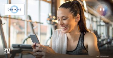 Junge Frau mit Handtuch über der Schulter sitzt im Fitnessstudio und lächelt beim Blick auf ihr Smartphone, während sie den digitalen Service MYCOACH von DSB-ONE nutzt.