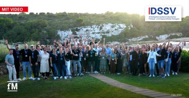 Eine große Gruppe von Menschen steht im Freien und posiert lachend für ein Gruppenfoto vor einer Küstenlandschaft mit Felsen und grünen Bäumen.