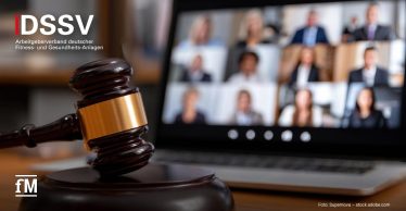 Ein Richterhammer liegt im Vordergrund auf einem Tisch. Im Hintergrund ist unscharf ein Laptop mit einer Videokonferenz zu sehen, in der mehrere Personen zugeschaltet sind.