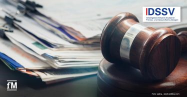 Im rechten Vordergrund liegt ein Holzrichterhammer auf einem runden Holzsockel auf einem dunklen Schreibtisch. Im linken Hintergrund befindet sich ein unscharfer Stapel mehrerer Papierakten mit unterschiedlich farbigen Trennblättern