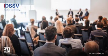 Konferenzraum aus hinterer Zuschauerperspektive; im Vordergrund mehrere sitzende Zuhörer auf Reihenstühlen mit Blick nach vorn, im Hintergrund Podium mit vier diskutierenden Personen vor Projektionsfläche