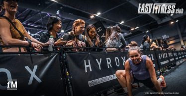 Zuschauende feuern in einer Messehalle eine HYROX-Athletin vor EASYFITNESS- und HYROX-Banden während eines Fitnessrennens an. Logos der beiden Marken sind auf der Bande und rechts oben.