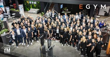 Große Gruppe von Mitarbeitenden in schwarzer oder dunkler Kleidung posiert auf einem Messestand mit Fitnessgeräten; oben steht groß „EGYM“.