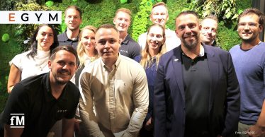 Eine Gruppe von Frauen und Männern steht vor einer begrünten Wand mit dem Schriftzug 'EGYM' und blickt in die Kamera.