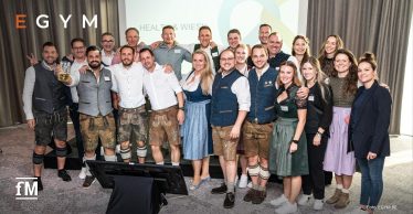 Eine große Gruppe von Personen in Trachtenkleidung steht vor einer Leinwand und lächelt in die Kamera.