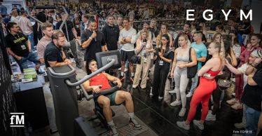 Messehalle mit EGYM-Fitnessgeräten, eine Person trainiert an einer Maschine, viele Menschen stehen um das Gerät herum und beobachten.