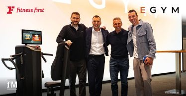 Im Vordergrund stehen vier Männer frontal neben einer schwarzen EGYM-Trainingsmaschine mit Sitz und Display: von links Daniel Kraus, Jan Scherzer, Martin Seibold und Dominik Klein. Links oben das rote Logo von Fitness First, rechts oben in der Ecke das von EGYM in Großbuchstaben, das E orangefarben, der Rest schwarz.