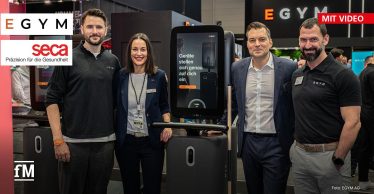 Vier Personen, darunter Mitarbeitende von EGYM und seca, stehen nebeneinander auf der Fitness-Messe FIBO vor einem EGYM-Gerät mit großem Display. Auf dem Bildschirm steht: „Geräte stellen sich genau auf dich ein“. Im Hintergrund sind weitere Messestände und Besucherinnen udn Besucher zu sehen. Links oben befinden sich die Logos von EGYM und seca.