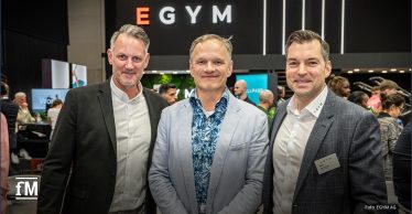 Drei Männer (von links Stefan Küper, Dr. Frank Schifferdecker-Hoch und Jan Scherzer) stehen nebeneinander und blicken lächelnd in die Kamera. Sie befinden sich auf einer gut besuchten Messe oder Veranstaltung, im Hintergrund ist deutlich der Schriftzug 'EGYM' auf einem schwarzen Messestand zu erkennen.