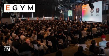 Große Bühne mit Projektionswand „The EGYM Journey“, zwei Sprecher stehen vorn, Publikum sitzt dicht in Reihen, Aufnahme von hinten über Zuschauer hinweg.