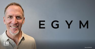Porträt eines lächelnden Mannes (Michael Baderenko) vor grauem Hintergrund. Rechts daneben steht in großen schwarzen Buchstaben „EGYM“.
