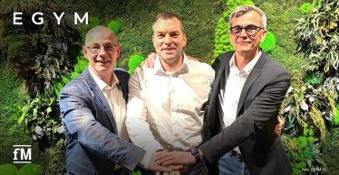 Drei Männer stehen frontal vor einer begrünten Wand mit großen weißen Buchstaben EGYM im oberen Hintergrund, ihre Hände liegen übereinander im Vordergrund, seitliche Pflanzenflächen rahmen die Szene ein.