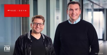 In einem hellen Büroflur mit Glaswänden stehen zwei Männer nebeneinander: Links steht ein Mann mit Brille (Philipp Roesch-Schlanderer) und dunkler Jacke, rechts ein Mann mit dunklem Pullover und beiger Hose (Fritz Lanman).