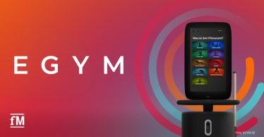 Großes weißes EGYM-Logo auf orange-rot-violettem Farbverlauf. Rechts steht ein Trainingsgerät mit integriertem Bildschirm.