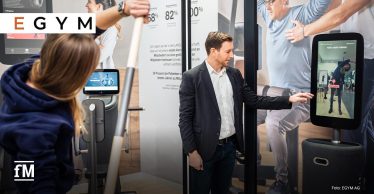 Ein EGYM-Mitarbeiter im Anzug steht auf einer Messe vor einem digitalen EGYM Gerät mit Bildschirm und zeigt mit dem Finger auf die Anzeige. Auf dem Bildschirm ist eine Frau zu sehen, die eine Übung ausführt. Im Vordergrund des Bildes übt die Frau mit einem Gymnastikstab. Im Hintergrund sind weitere Trainingsgeräte sowie großformatige Plakate mit erklärenden Texten und Bildern zu sehen.