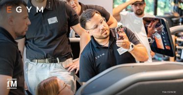 Mehrere Personen in schwarzer EGYM-Kleidung betrachten gemeinsam ein Trainingsgerät mit Bildschirm; ein Mann hält ein Smartphone mit geöffneter App in der Hand.