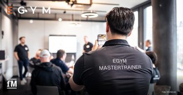 Eine Person mit 'EGYM MASTERTRAINER'-Shirt filmt eine Präsentation in einem modernen Schulungsraum mit mehreren Teilnehmenden und Vortragenden.