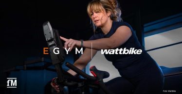 Frau trainiert auf einem Wattbike mit Display; Logos von EGYM und Wattbike im Vordergrund
