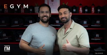 Zwei Männer stehen lächelnd am Service-Point eines Fitnessstudios und zeigen den Daumen nach oben, im Hintergrund Regale mit Trinkflaschen.