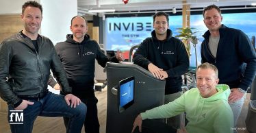 Fünf Männer (Markus Hangl (INVIBE), Hannes Götze (Wellpass), Kim Fischer (Wellpass), Philip Zischke (INVIBE) stehen in einem modernen Fitnessstudio rund um ein Trainingsgerät von EGYM. Florian Streiter (INVIBE) sitzt auf dem Gerät, lächelt und zeigt den Daumen nach oben