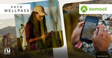 Person mit Rucksack nutzt ein Smartphone im Freien; daneben zeigt ein Smartphone die komoot-App mit Routenkarte, EGYM-Wellpass- und komoot-Logos sichtbar